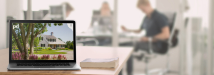 Laptop on a wooden desk displays a photo of a white house with a green lawn and trees; a blurred office with people sits in the background.