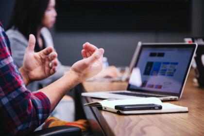 Two people in a meeting; one person gestures with open hands while a laptop, notebook, and phone sit on the table.