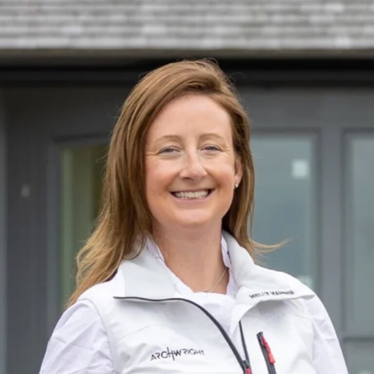 Smiling person with light brown hair wearing a white jacket with a small logo, standing outdoors in front of a gray building.