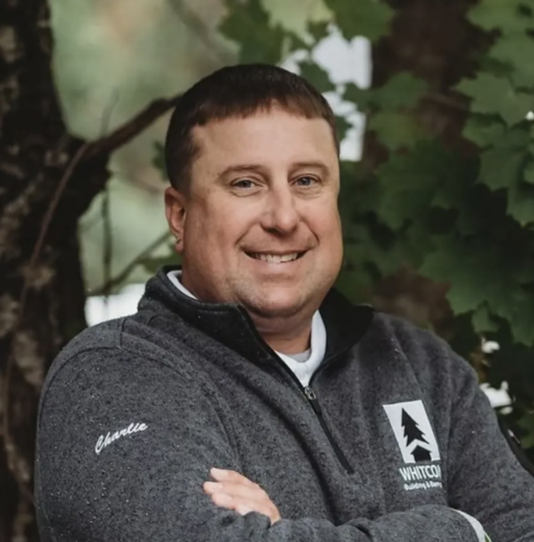 Smiling person with short brown hair, wearing a dark gray zip-up sweater with a white chest logo and a sleeve patch, arms crossed, outdoors among greenery.