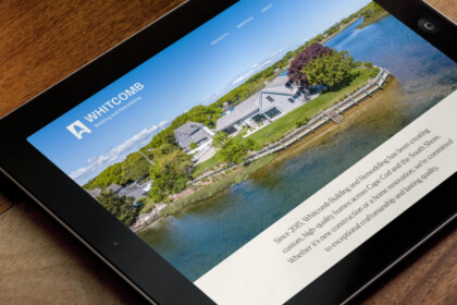 Tablet on a wooden desk displaying the Whitcomb Building and Remodeling site, featuring a waterfront home, dock, trees, and blue sky.