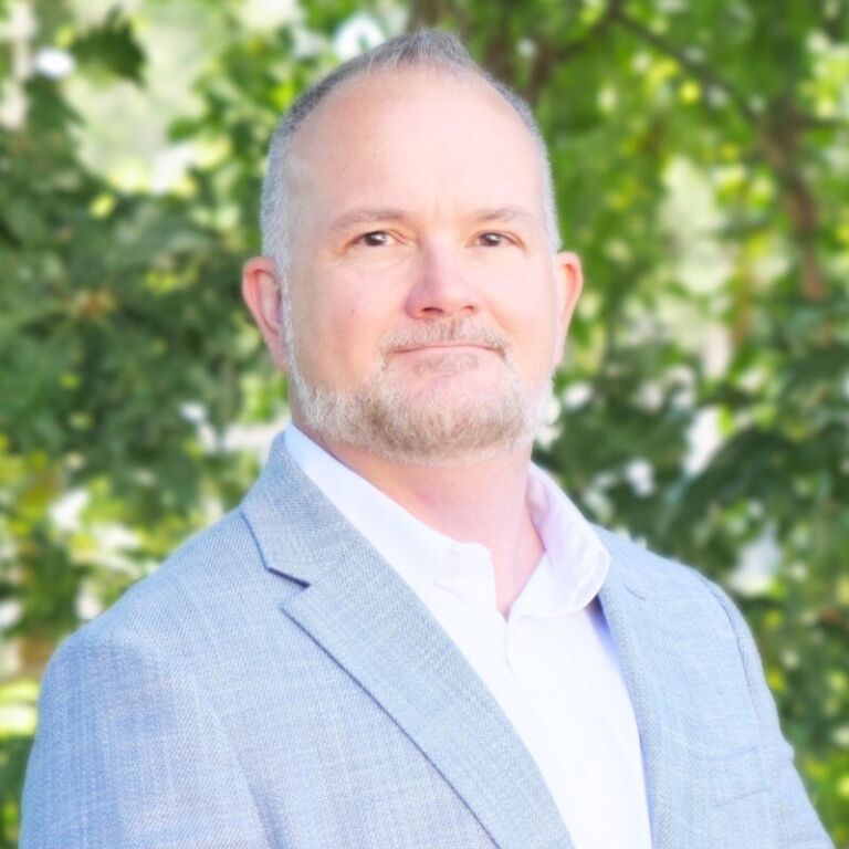 Portrait of a person with short gray hair and a trimmed beard, wearing a light blue blazer and white shirt, outdoors with green foliage in the background.