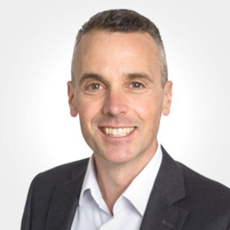 Head-and-shoulders portrait of a person with short dark hair, wearing a white shirt and dark blazer, smiling against a light neutral background.