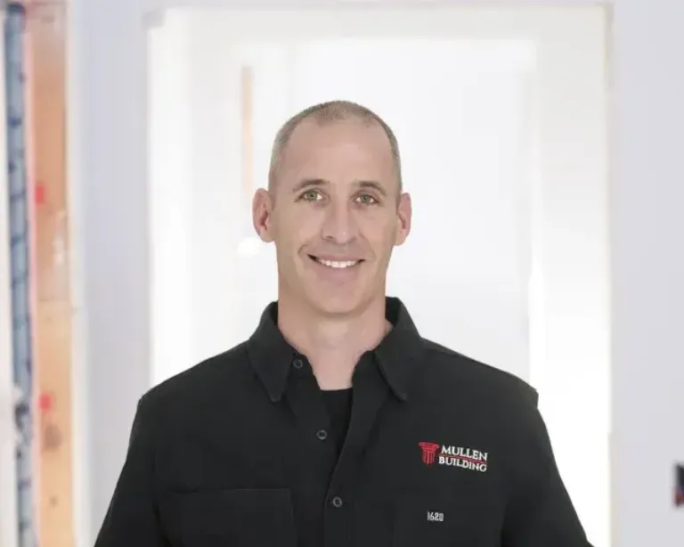 A person with short hair wearing a black polo shirt with a red-and-white Mullen Building logo, smiling in a bright, well-lit hallway.