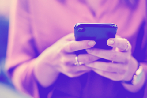 A person holding a blue smartphone with both hands, wearing rings and a wristwatch, against a purple, blurred background.