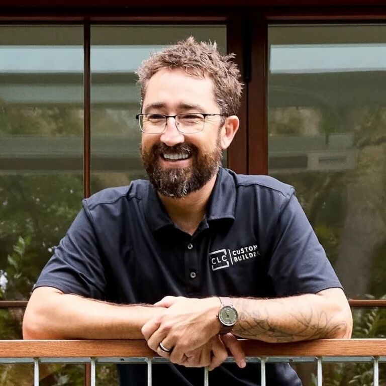 A smiling person with short gray hair and a beard, wearing glasses and a dark polo with a "Custom Builders" logo, leaning on a wooden railing outdoors.