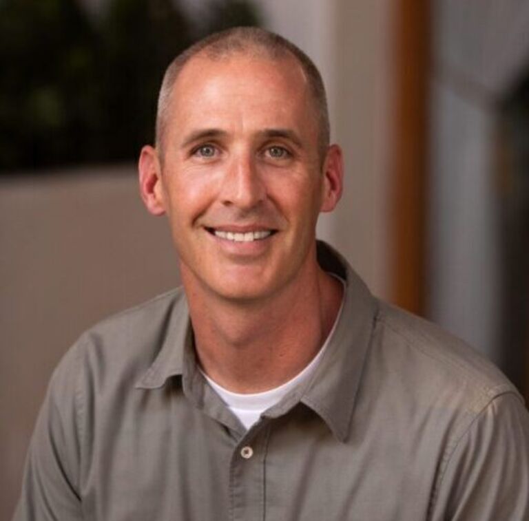 Portrait of a person with short light hair, smiling warmly, wearing a light gray button-up shirt over a white tee, indoors.