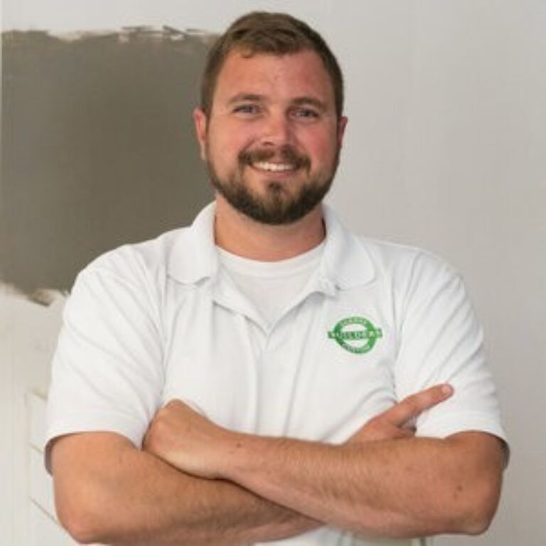 A smiling person with short hair and a beard stands with arms crossed, wearing a white polo shirt featuring a green circular logo on the chest.