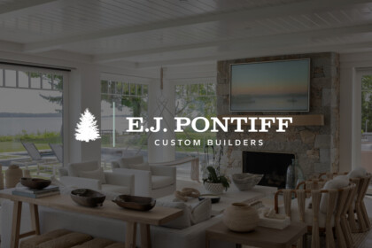 Bright, airy living room with white furniture, stone fireplace, large windows over a lake, and a mounted TV; overlaid logo text: “E.J. Pontiff Custom Builders” with a pine tree icon.