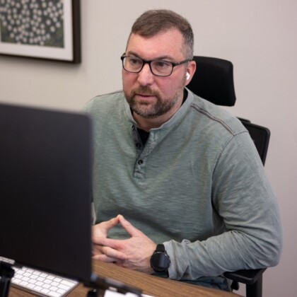 Man with short gray hair and beard, wearing glasses and a light green henley, sits at a desk in front of a computer with earbuds in, hands clasped.