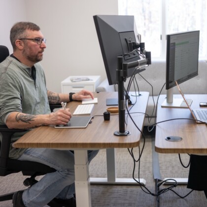 A man with tattoos and glasses sits at a wooden desk with a large monitor, stylus tablet, and keyboard, working in a bright office.