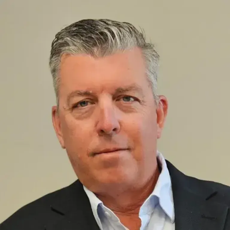 Close-up portrait of a middle-aged person with short gray hair, wearing a light blue shirt and dark blazer, neutral expression, neutral beige background.