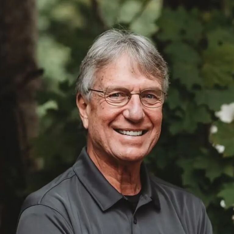 Smiling older person with short gray hair and glasses wearing a dark shirt, outdoors with green foliage in the background.