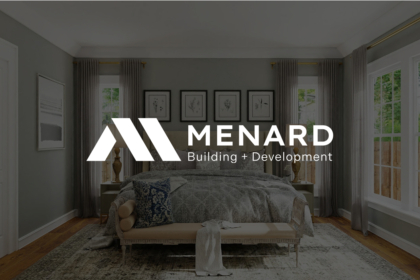 Interior view of a stylish bedroom with a large bed, gray walls, framed art above, tall windows with drapes, and a prominent white “MENARD Building + Development” logo overlay.
