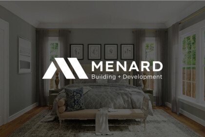 Bedroom interior with neutral gray walls, large windows, sheer curtains, and a plush bed; overlaid white Menard Building + Development logo in center.
