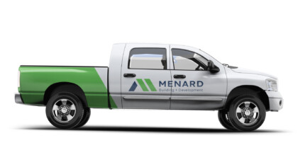 Pickup truck with white cab and green rear, side logo "MENARD Building + Development" in blue and green, black tires, chrome trim, company branding.