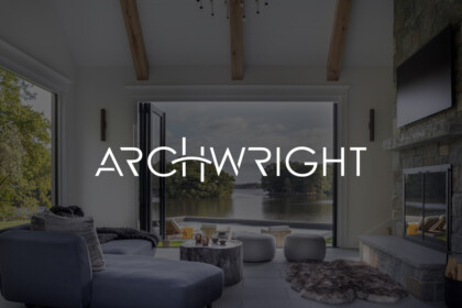 Living room with large open doors leading to a waterfront view, modern gray sectional, stone fireplace, wooden ceiling beams, and outdoor seating by the water.