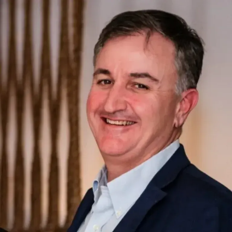 Smiling man with short dark hair in a dark blazer and light blue shirt, standing indoors with curtains in the background.