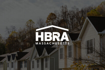 Neighborhood row of pastel townhouses with autumn trees in background, overlaid by a bold HBRA Massachusetts logo in white.