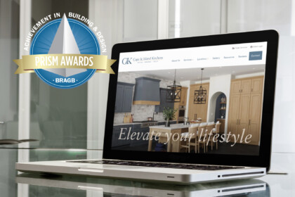 Laptop displaying Cape & Island Kitchens website with "Elevate your lifestyle" tagline, alongside a BRAGB Prism Awards badge for Achievement in Building & Design, positioned on a reflective glass surface with windows in the background.