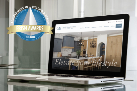 Laptop displaying Cape & Island Kitchens website with "Elevate your lifestyle" tagline, alongside a BRAGB Prism Awards badge for Achievement in Building & Design, positioned on a reflective glass surface with windows in the background.